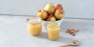 Auf dem Bild sieht man in Gläser abgefülltes Apfelmus. Daneben ein paar Äpfel und Zimtstangen. Apfelmus