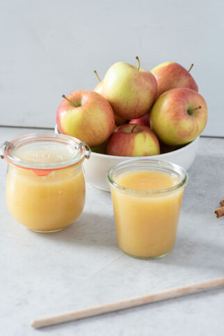 Auf dem Bild sieht man in Gläser abgefülltes Apfelmus. Daneben ein paar Äpfel und Zimtstangen. Apfelmus
