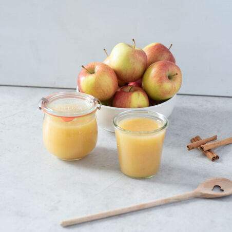 Auf dem Bild sieht man in Gläser abgefülltes Apfelmus. Daneben ein paar Äpfel und Zimtstangen. Apfelmus